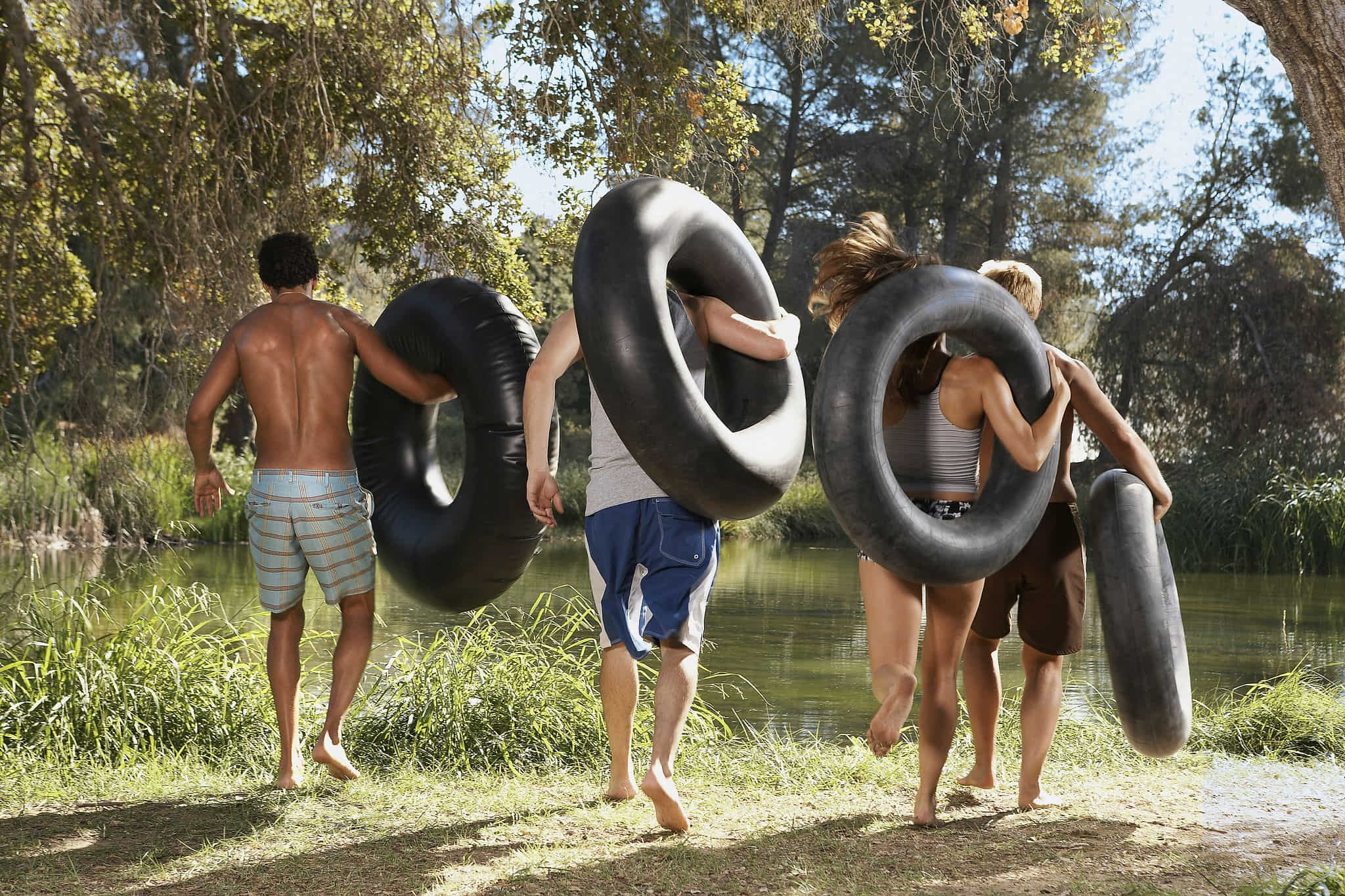 Four young adults carrying large black inner tubes running toward a calm green river through a sun-dappled forest bank on a bright summer afternoon — book a fee-free water tubing vacation rental on Houfy | Houfy — No Fees, Book Direct
