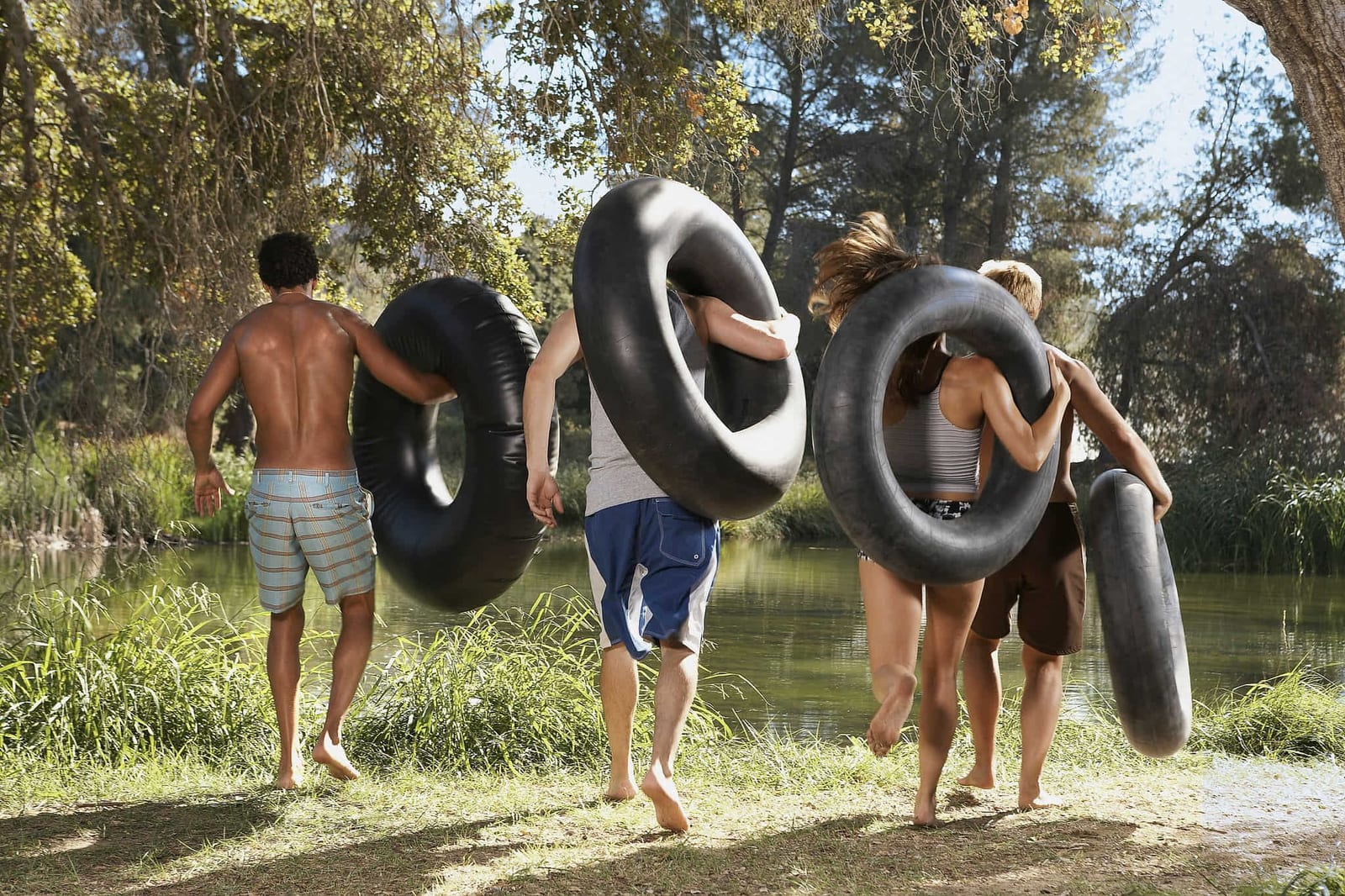 Four young adults carrying large black inner tubes running toward a calm green river through a sun-dappled forest bank on a bright summer afternoon — book a fee-free water tubing vacation rental on Houfy | Houfy — No Fees, Book Direct