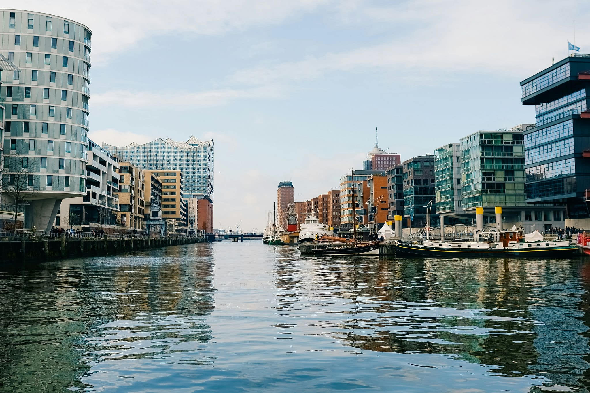 Serene waterfront canal view in Hamburg's HafenCity district, with the iconic wave-like roofline of the Elbphilharmonie, modern glass apartment balconies, moored sailing vessels, and rippling teal water reflecting the hazy sky — book a fee-free water view vacation rental on Houfy | Houfy — No Fees, Book Direct