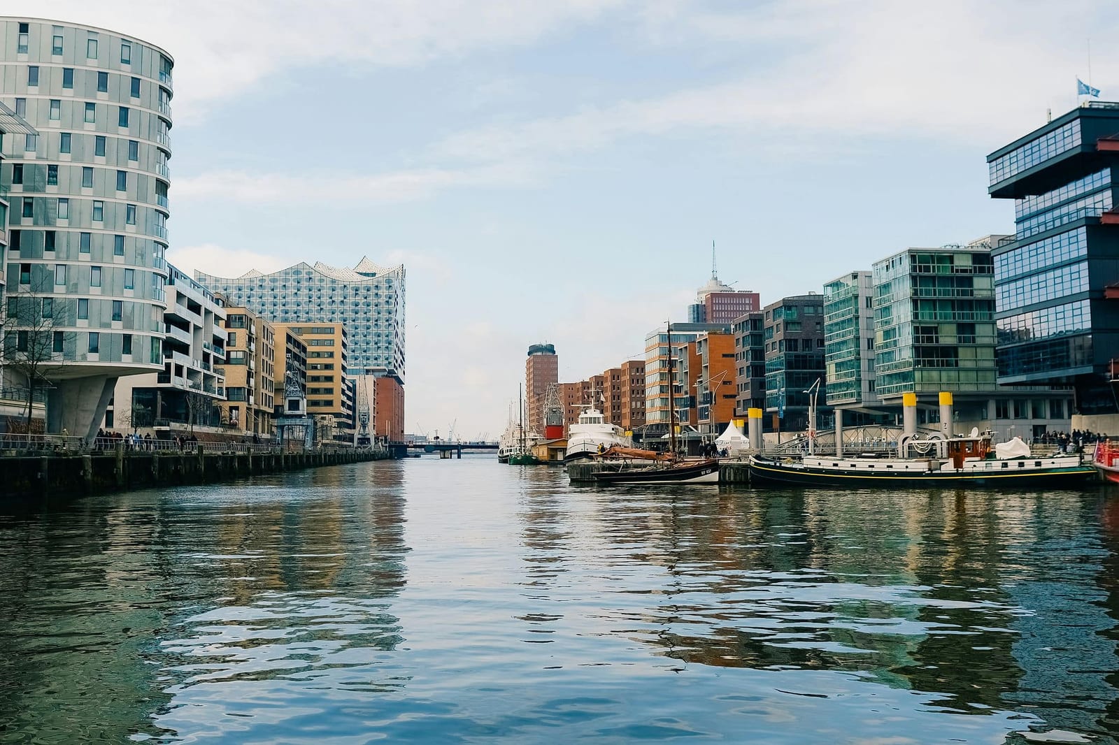 Serene waterfront canal view in Hamburg's HafenCity district, with the iconic wave-like roofline of the Elbphilharmonie, modern glass apartment balconies, moored sailing vessels, and rippling teal water reflecting the hazy sky — book a fee-free water view vacation rental on Houfy | Houfy — No Fees, Book Direct