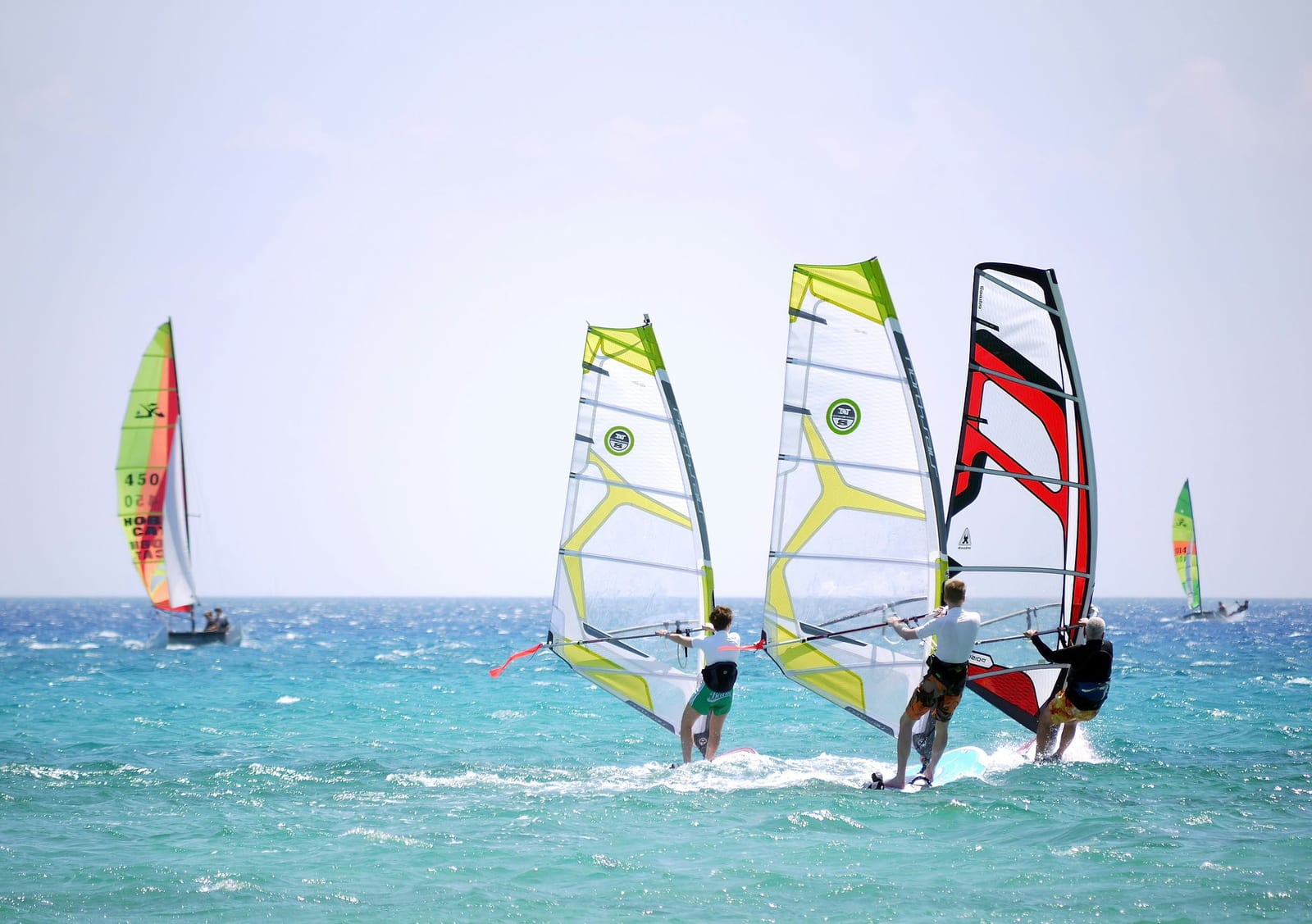 Three windsurfers with lime green and red North Sails racing across brilliant turquoise ocean water with white foam spray, leaning into a steady wind under a bright midday sky, with a Hobie Cat catamaran sailing in the background — book a fee-free windsurfing vacation rental on Houfy | Houfy — No Fees, Book Direct