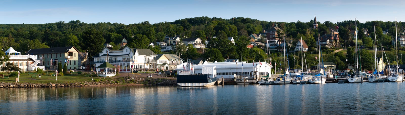 Bayfield harbor with moored sailboats and charming houses on hilly terrain, surrounded by lush greenery. Historic buildings dot the landscape, offering a picturesque view unique to Wisconsin's Lake Superior shoreline. | Houfy — No Fees, Book Direct