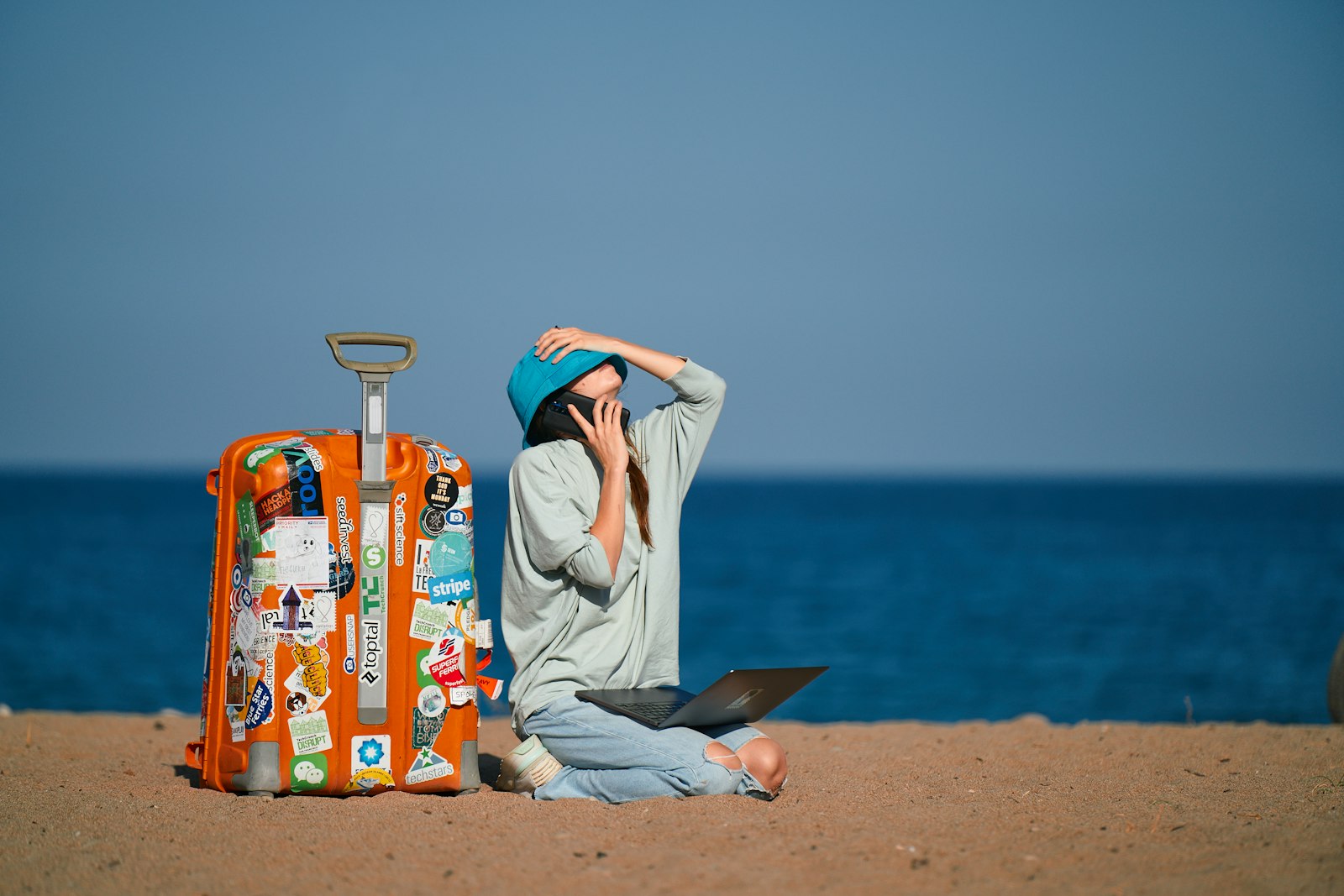 A traveler sitting on a beach with a laptop and a suitcase, planning a trip