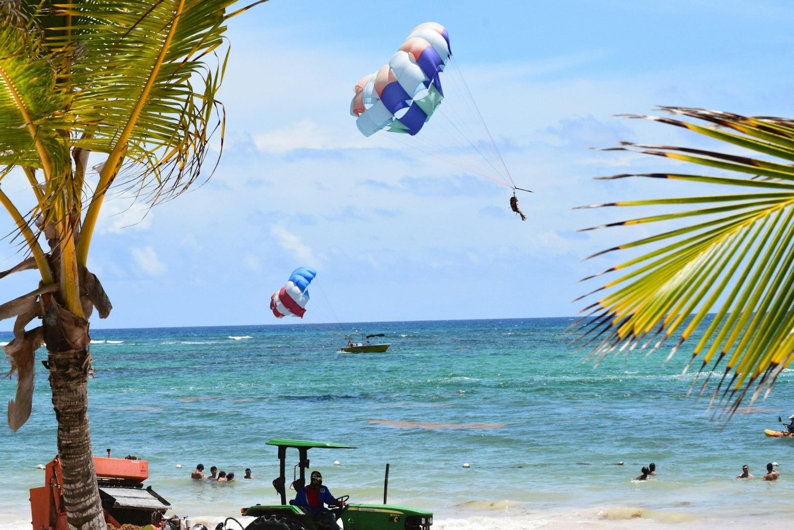 Parasail rising over turquoise Caribbean water off a busy beach, with boats and swimmers below.