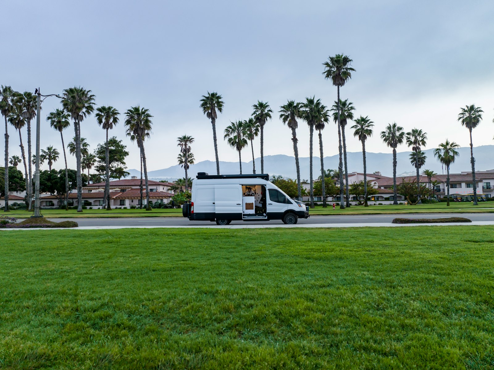 White passenger van on a palm-lined road under a bright sky.