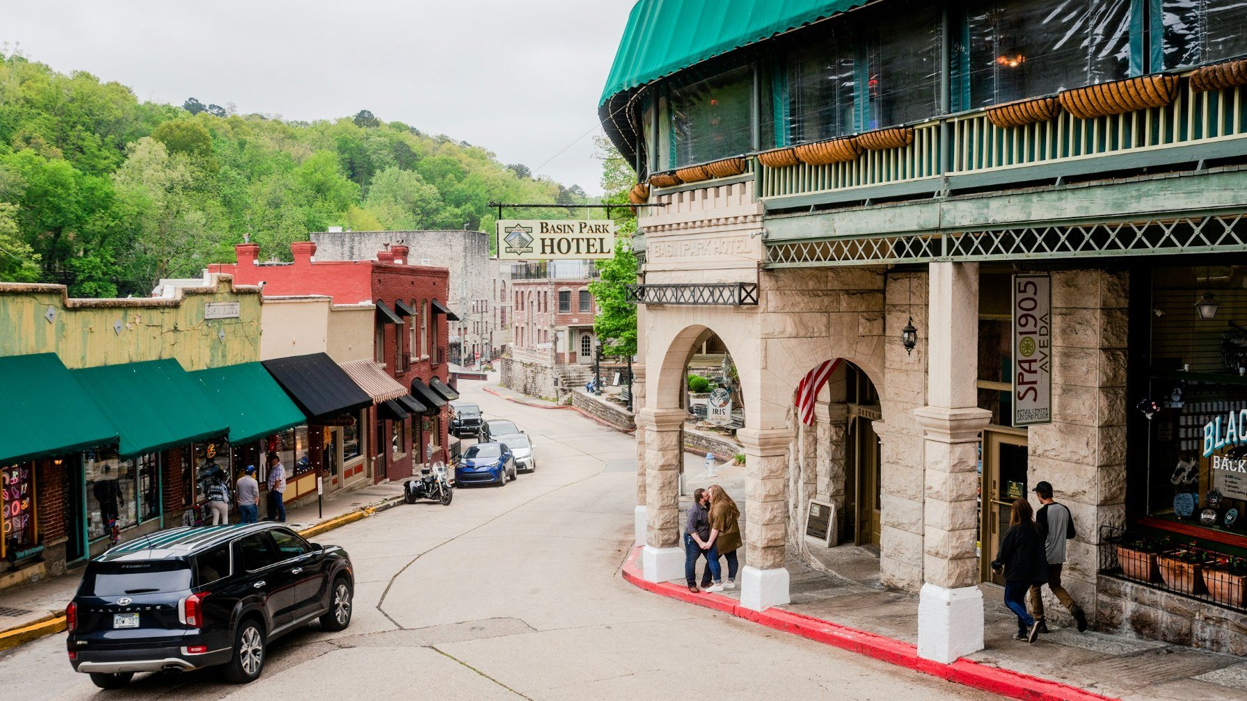 A hillside street of stone and brick buildings in downtown Eureka Springs, with a Basin Park Hotel sign and green wooded hills behind