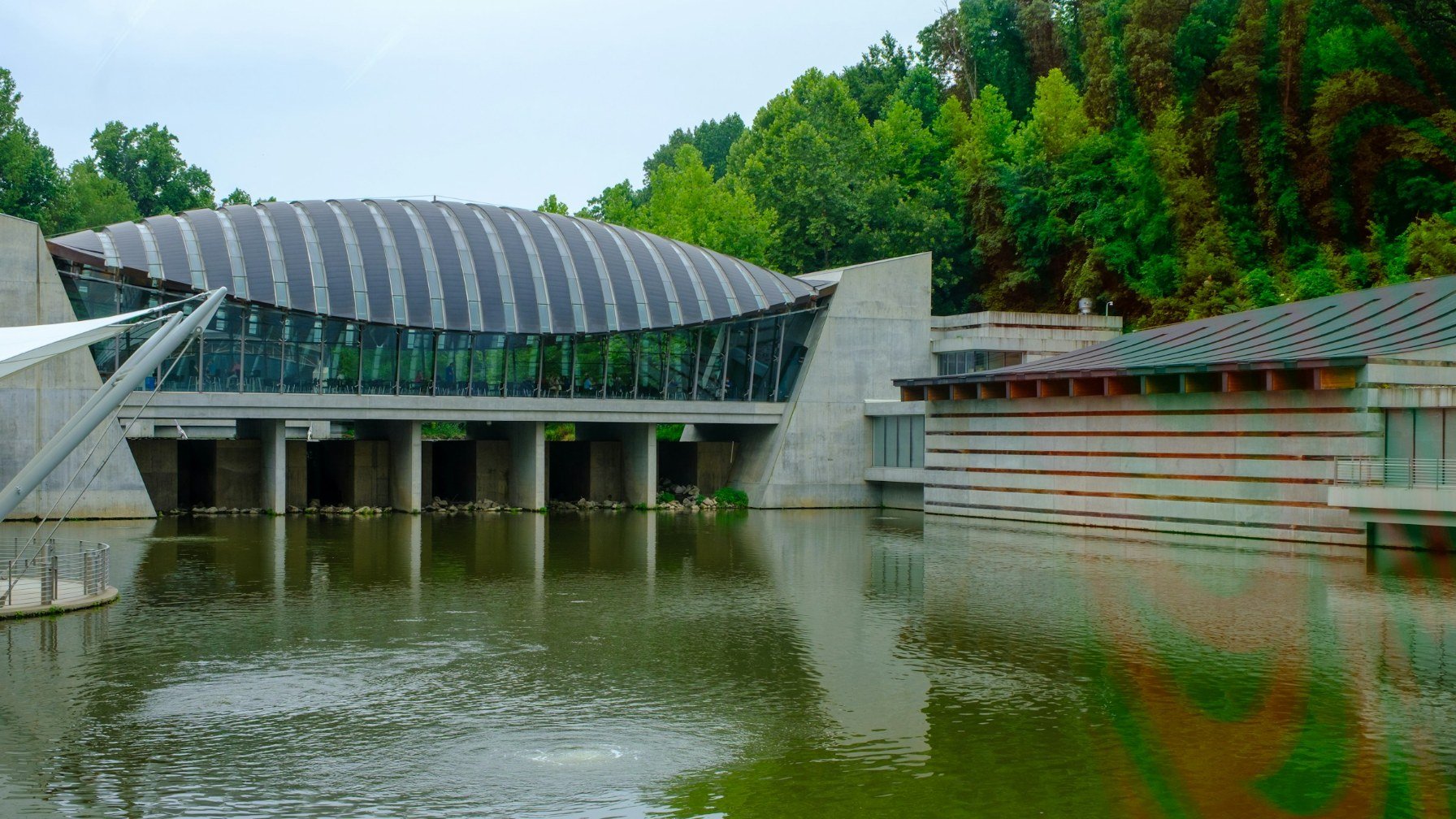 Curved metal roof of the Crystal Bridges Museum of American Art spanning a pond in Bentonville, Arkansas, with wooded hillside behind