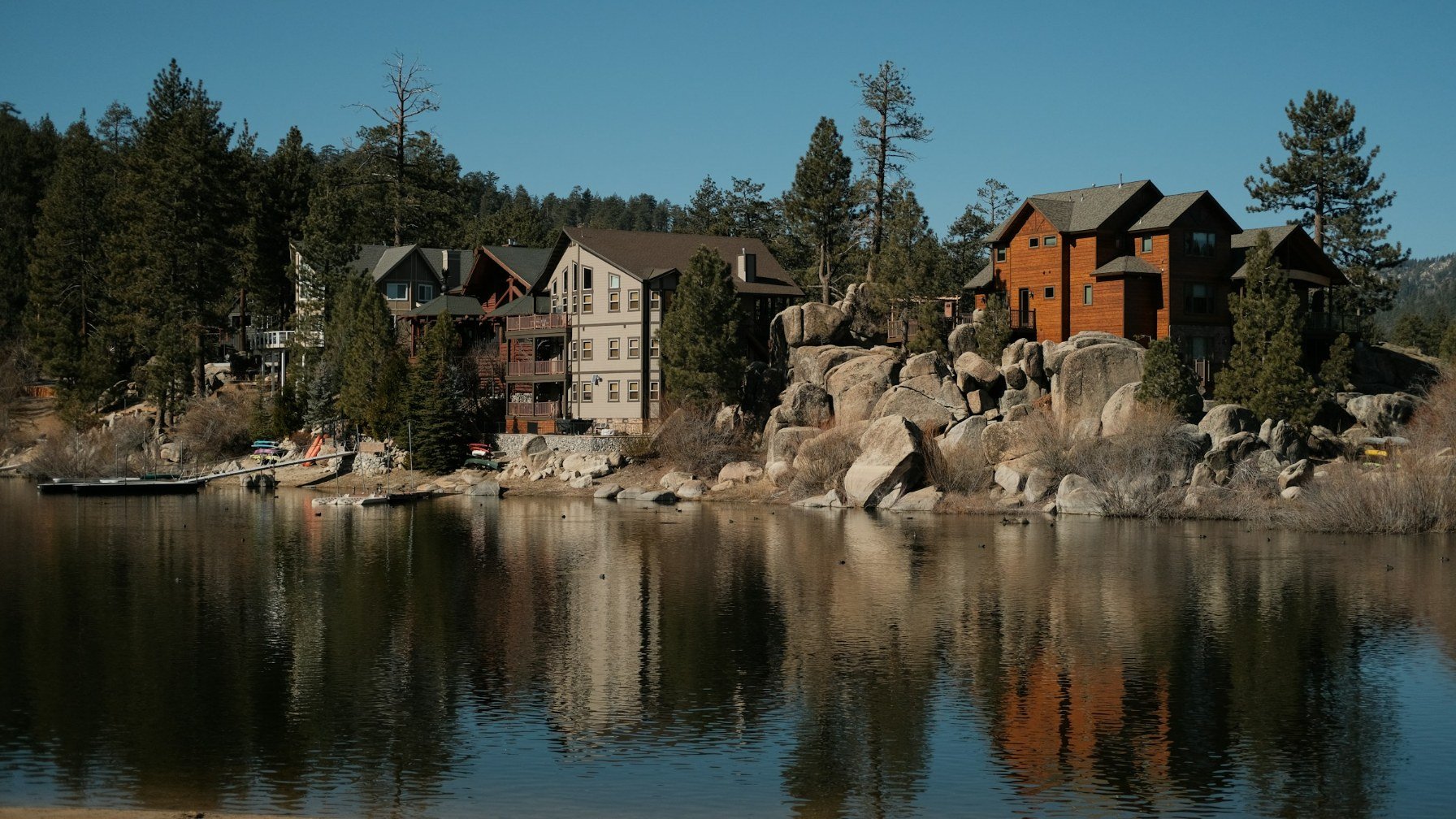 Wood-clad lakeside houses among large granite boulders and pine trees at Big Bear Lake, reflected in still water under a clear blue sky