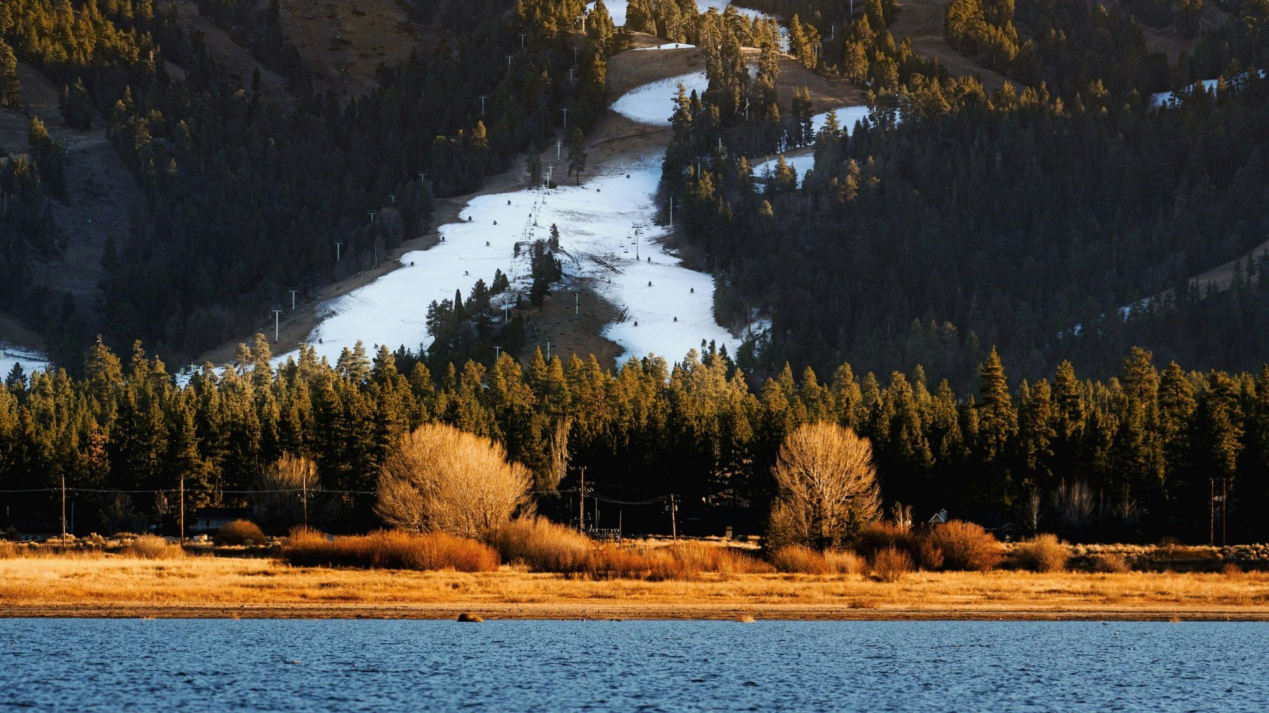 A ribbon of snow on a forested ski slope above a dark blue lake, with golden grass along the shoreline and a clear sky