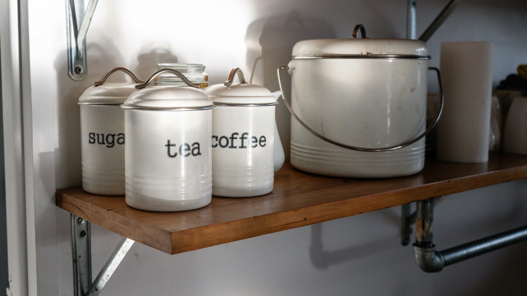 Three white canisters labelled sugar, tea and coffee on a wooden shelf beside a kettle