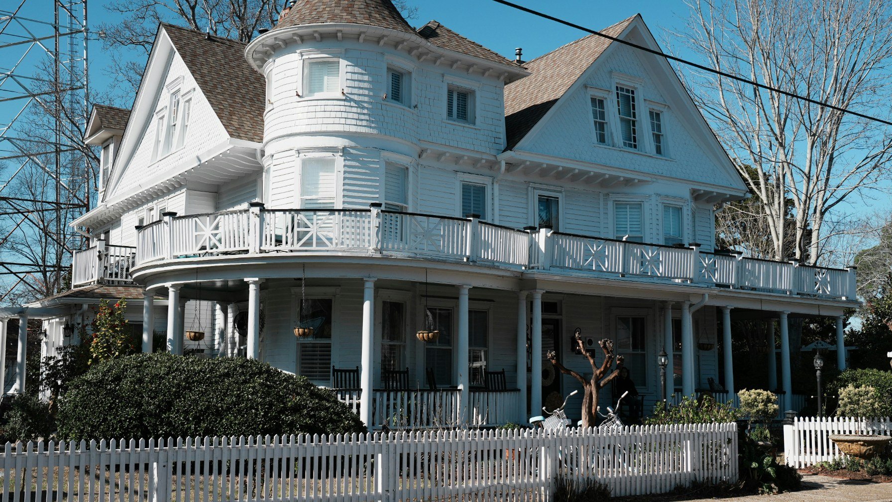 A large white Victorian house with a wraparound porch and a corner turret behind a picket fence
