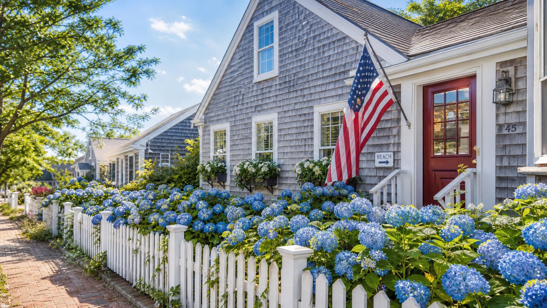 Cape Cod gray shingle beach cottage vacation rental with American flag at the front door and blue hydrangeas blooming along the white picket fence on a sunny July afternoon