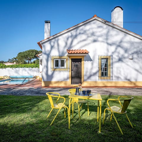 A bright, open garden space with sunlight bouncing off the white walls and a cosy table set in warm yellow tones, with the pool tucked to the far left.