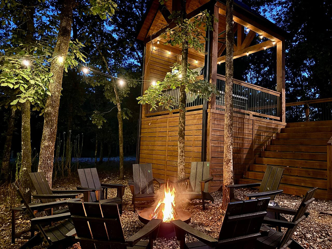 Cozy fire pit with treehouse and glowing string lights in forest setting.