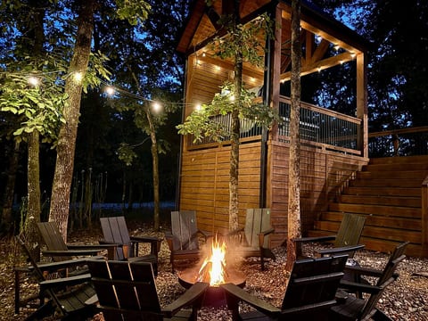 Cozy fire pit with treehouse and glowing string lights in forest setting.