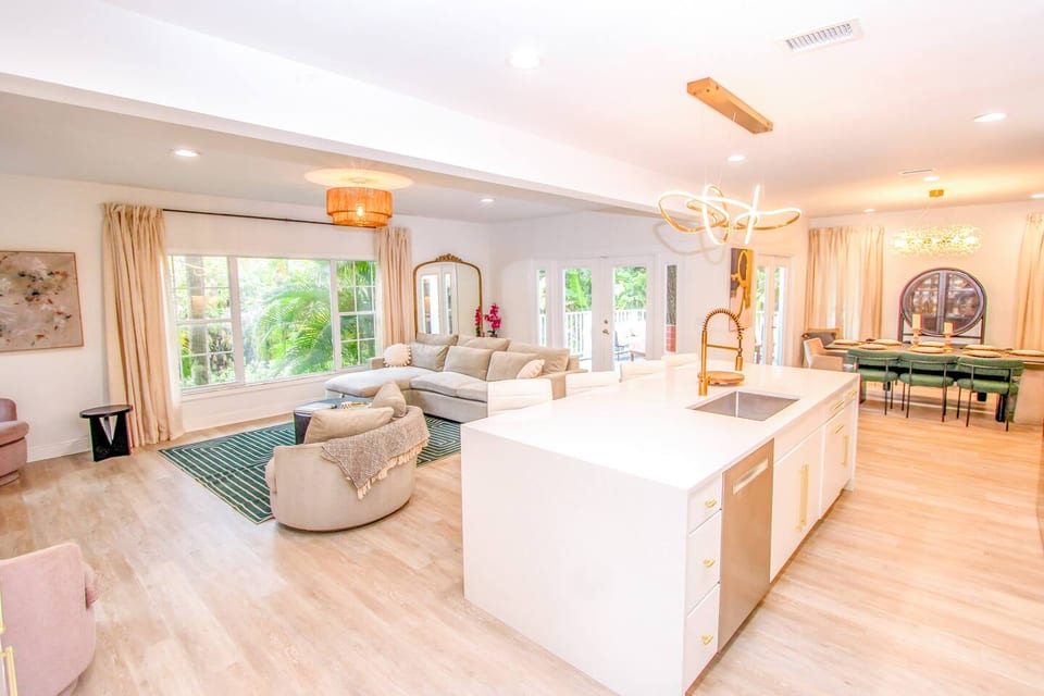 The open floor plan features an 8-foot island, connecting the gourmet kitchen, living, and dining areas. Surrounded by palm and oak trees, large windows offer views of nature w french doors leading to the outdoor deck and fully enclosed backyard 
