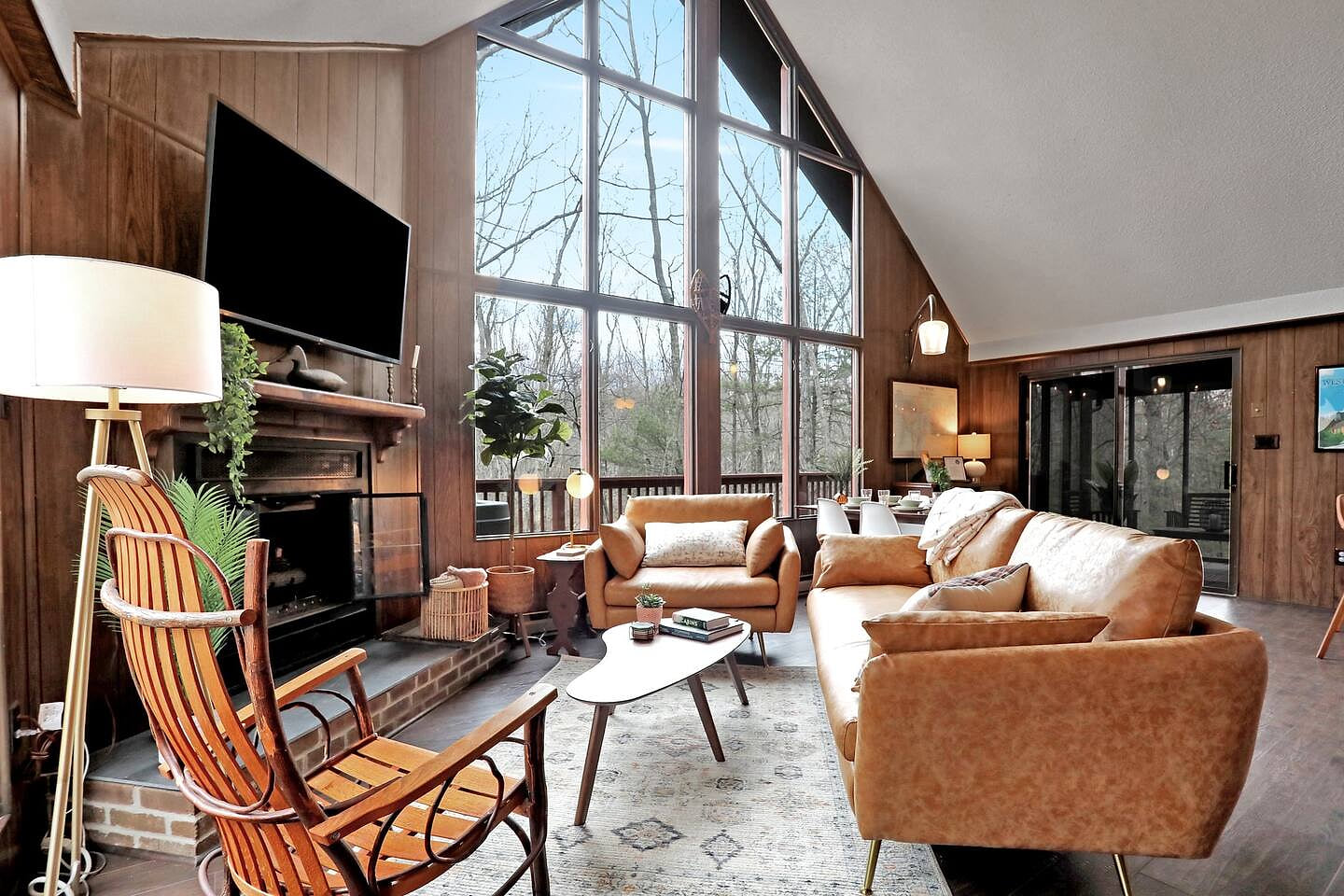 Stylish midcentury living room, dining area, and kitchen with huge windows facing the woods, high vaulted ceiling, and a gas fireplace.