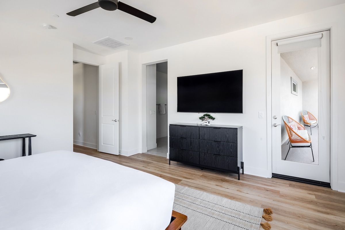 Enjoy a bright and modern bedroom featuring a large TV, private sauna, small desk for remote work, ensuite bathroom, and direct patio access with vibrant seating