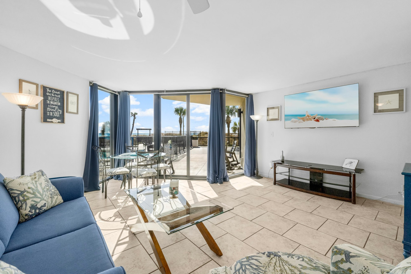 Bright and airy living space features a queen-size sleeper sofa, a mounted smart TV, and floor-to-ceiling windows with access to a private patio offering stunning pool and beach views.
