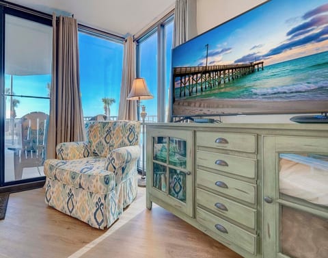 Coastal furnishings, a patterned armchair, television, and beachfront artwork create a welcoming corner beside the balcony doors.