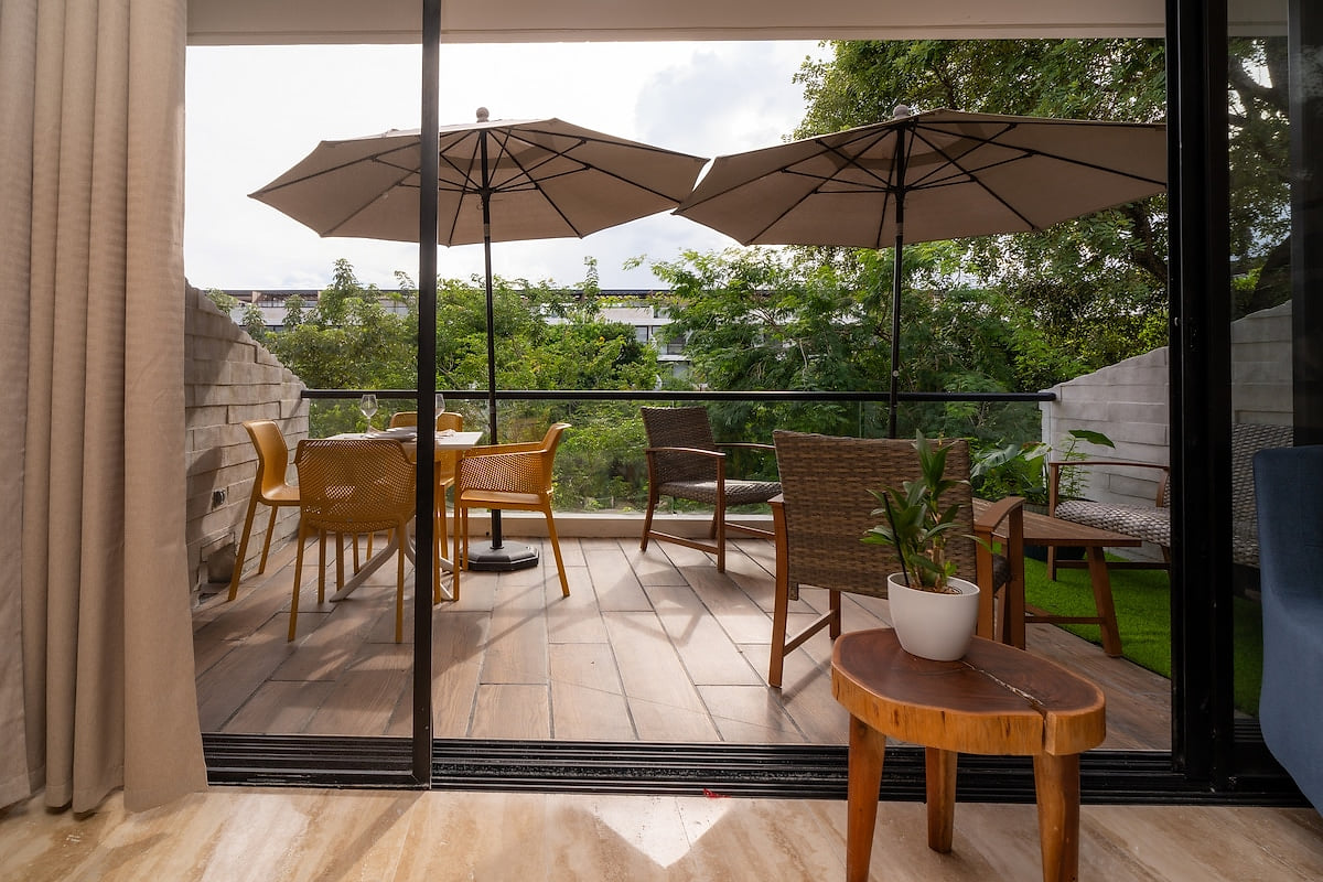 Serene patio with shaded dining area and comfortable seating. Lush greenery and sliding glass doors create a relaxing outdoor space.