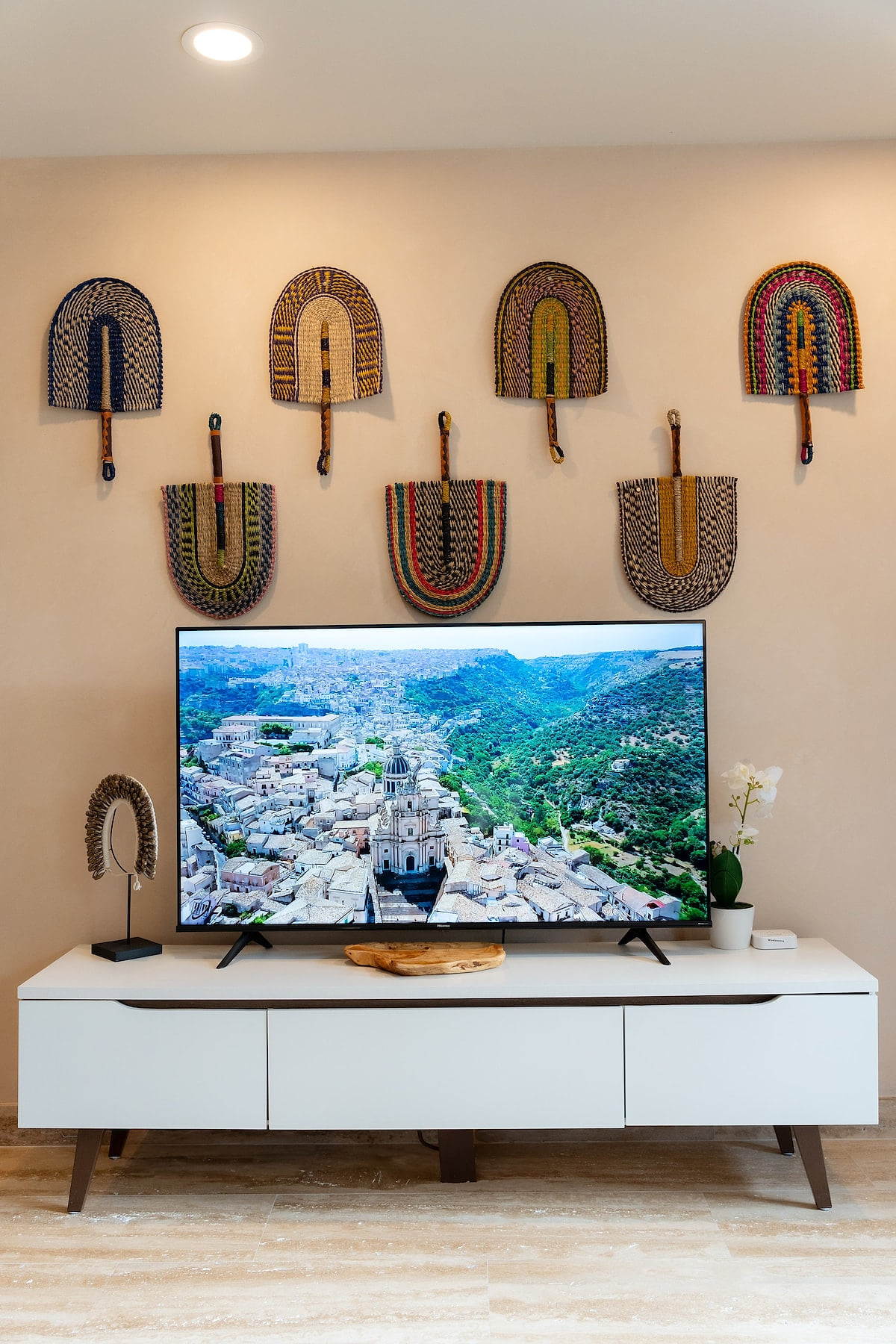Modern living room with wall decor of colorful fans. Large TV displays scenic image. Minimalist aesthetic with white TV stand and potted orchid.