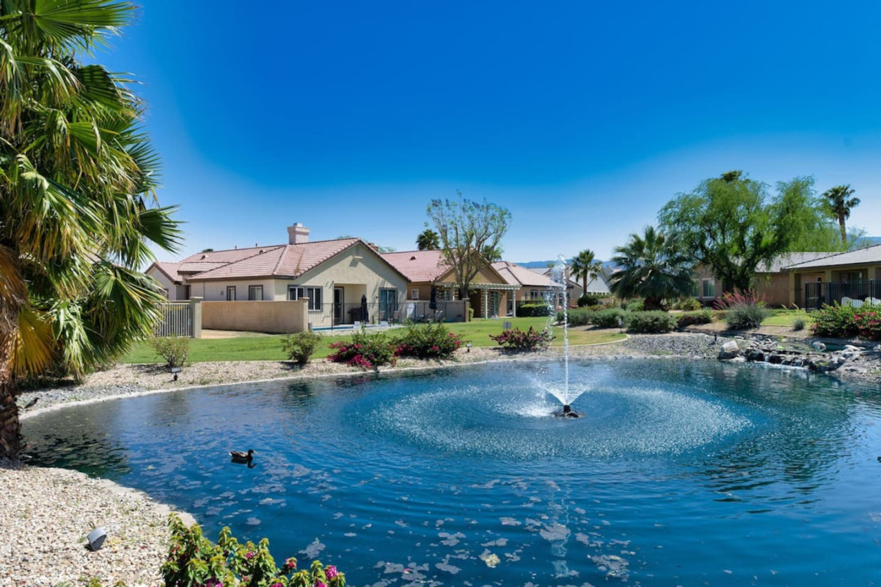 Peaceful pond w/ a bubbling fountain, blue skies, & landscaped surroundings for a calm outdoor backdrop.