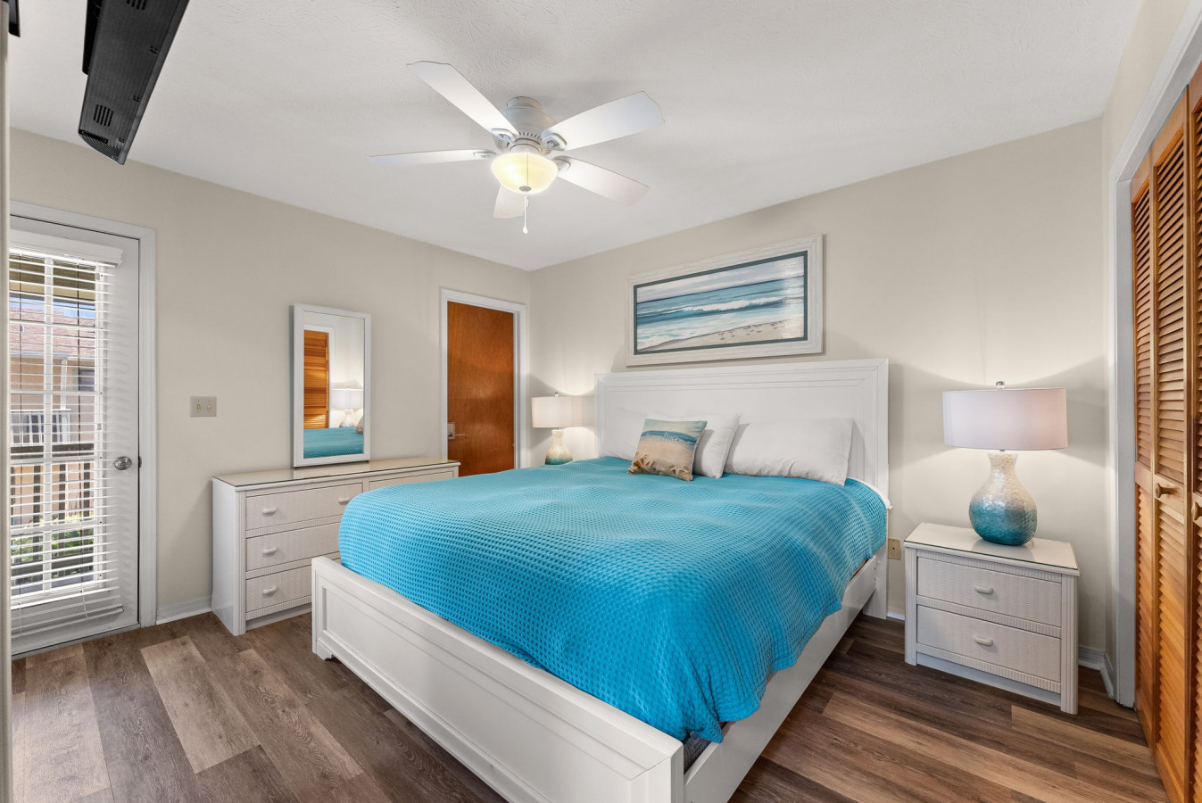 Bright coastal bedroom with a plush king bed, beach artwork, and private balcony access.