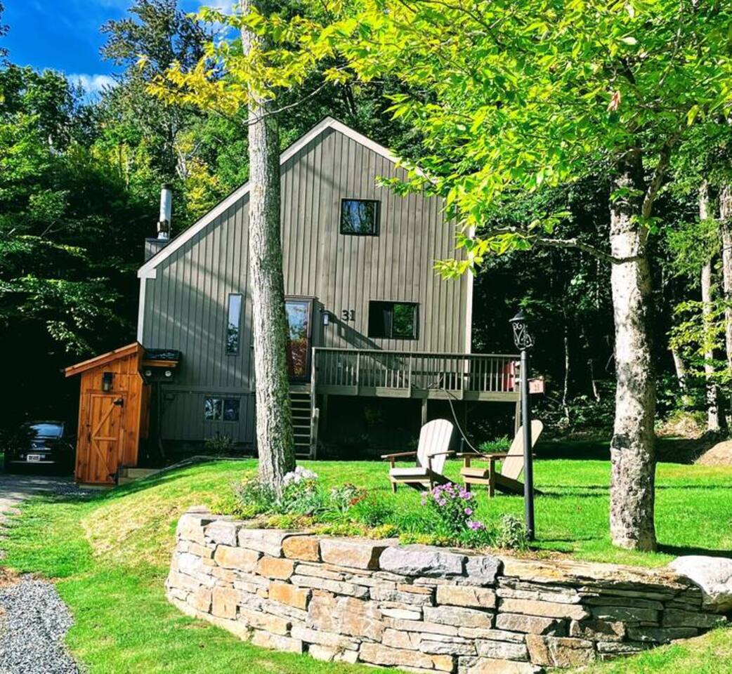 A cozy cabin-style home with a wooden deck, nestled among lush greenery, featuring Adirondack chairs on a stone-lined lawn.
