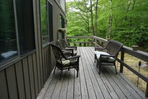 A rustic wooden deck extends from the house, surrounded by lush greenery. Three wicker chairs with cushions offer a peaceful spot to relax and enjoy the natural surroundings.