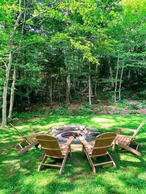 A cozy backyard fire pit area with four wooden Adirondack chairs arranged around a circular stone pit. Surrounded by lush green grass and dense trees, it offers a serene outdoor retreat.