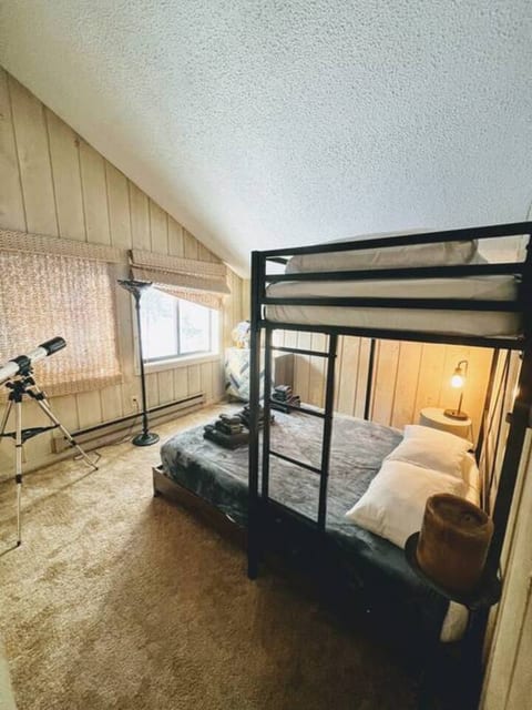 A cozy bedroom featuring a black metal bunk bed with a full-size lower and twin upper. The wooden-paneled walls and carpeted floor add warmth, while a telescope by the window invites stargazing.