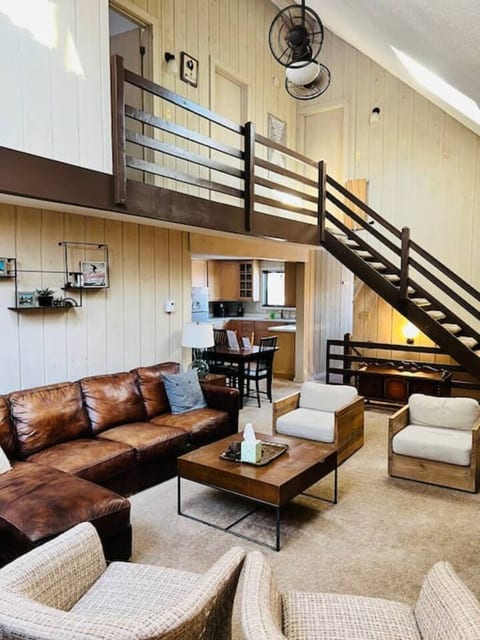 A cozy living room with a loft, featuring a leather sectional, woven chairs, and a rustic staircase leading to an upper level.