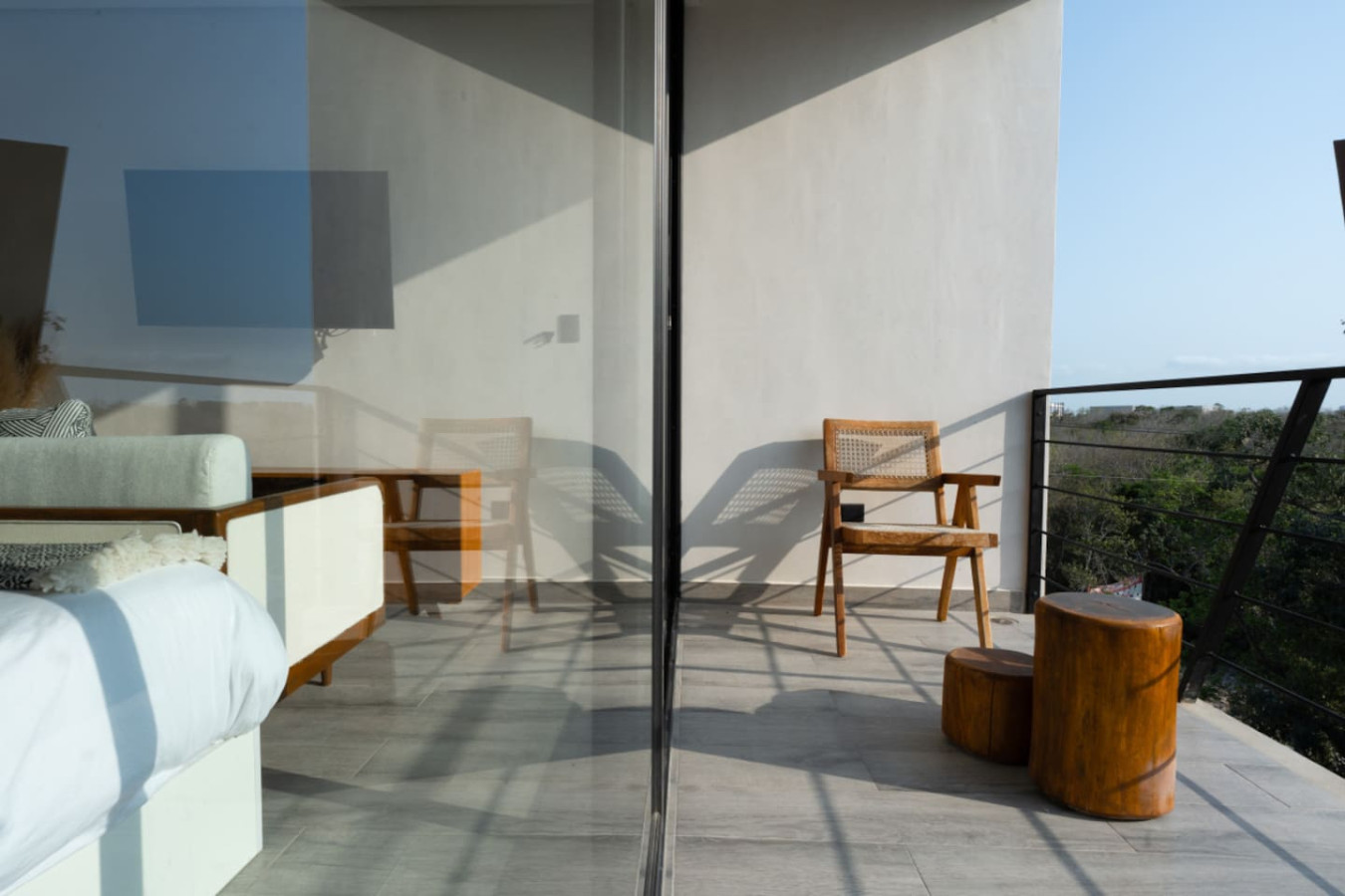 This minimalist balcony features two wooden chairs, natural wood stools, and glass sliding doors that reveal a cozy bedroom—nestled among treetops with open sky views.