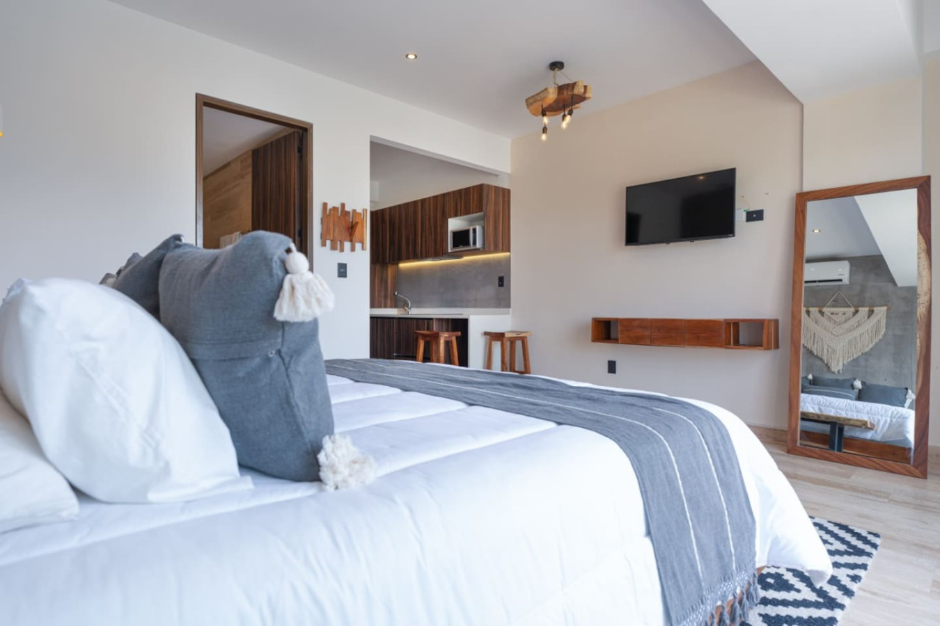 Modern suite with king bed, smart TV, full-length mirror, kitchenette, and bar seating. Natural wood accents and soft lighting create a warm, welcoming space.