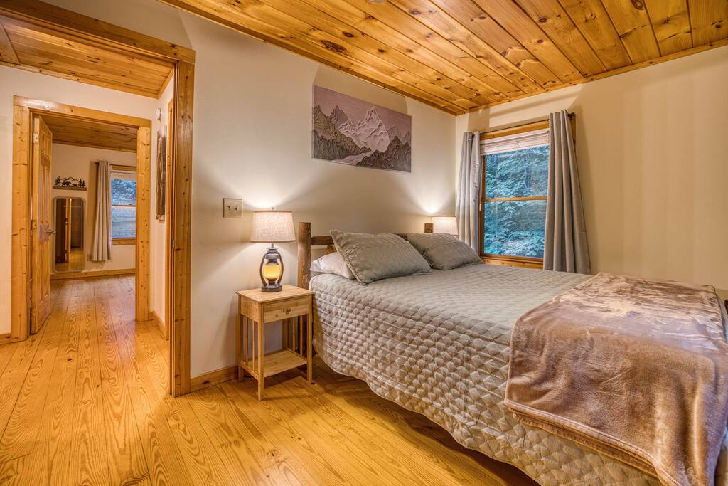 Cozy guest bedroom with a queen bed, plush bedding, and warm wood ceiling. Mountain art, bedside lamps, a scenic window with curtains, and a smart TV create a relaxing vibe. Cozy guest bedroom with a queen bed, plush bedding, and warm wood ceiling. Mountain art, bedside lamps, a scenic window with curtains, and a smart TV create a relaxing vibe. - Spacious Cabin, Hot Tub, Theater, Dog Run, FirePit - Ellijay