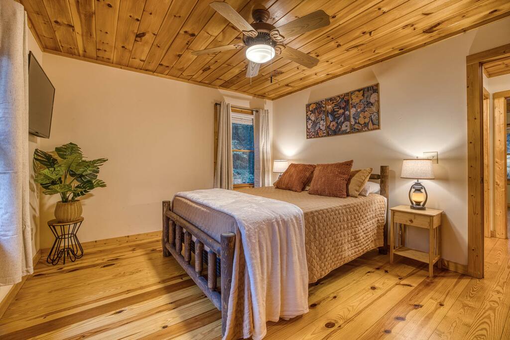Relax in the spacious guest bedroom featuring a queen-sized luxury mattress, rustic nightstands with lamps, a large closet, Smart TV, and a cozy rocking chair. The perfect blend of comfort and charm for a restful stay! Relax in the spacious guest bedroom featuring a queen-sized luxury mattress, rustic nightstands with lamps, a large closet, Smart TV, and a cozy rocking chair. The perfect blend of comfort and charm for a restful stay! - Spacious Cabin, Hot Tub, Theater, Dog Run, FirePit - Ellijay