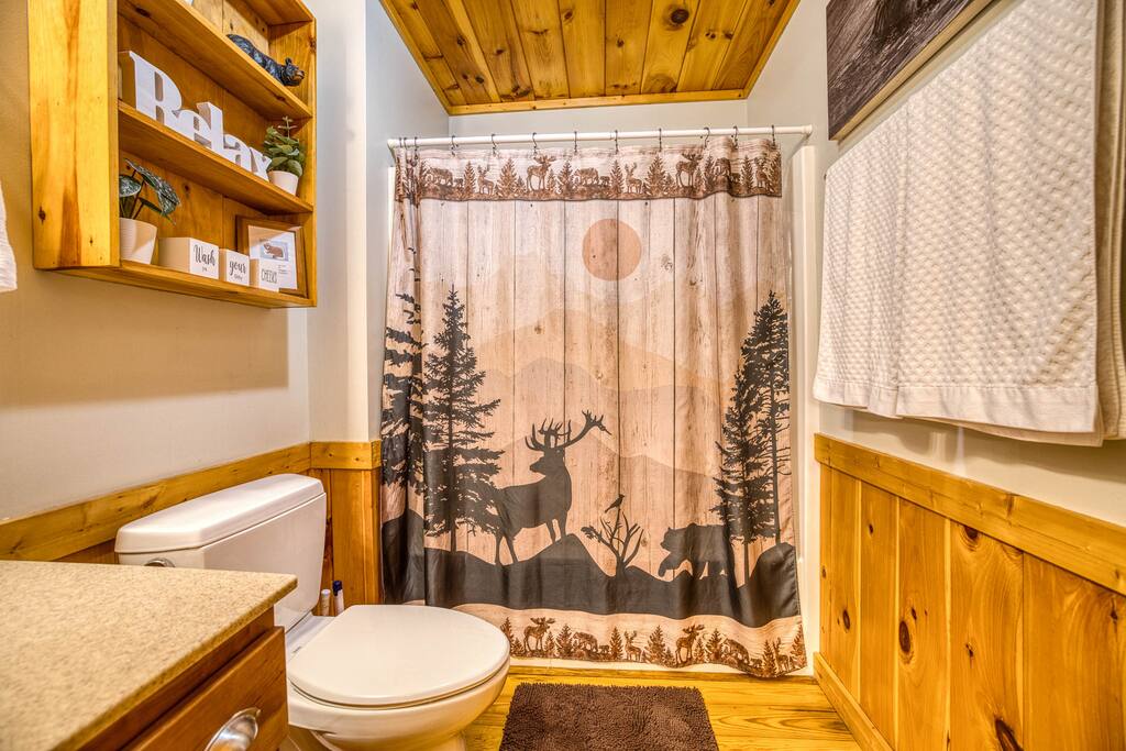 Cozy guest bathroom with a warm wood ceiling and wainscoting. Features a scenic shower curtain with deer and forest motifs, a single sink vanity, toilet, and shelving with plants and decor. Cozy guest bathroom with a warm wood ceiling and wainscoting. Features a scenic shower curtain with deer and forest motifs, a single sink vanity, toilet, and shelving with plants and decor. - Spacious Cabin, Hot Tub, Theater, Dog Run, FirePit - Ellijay