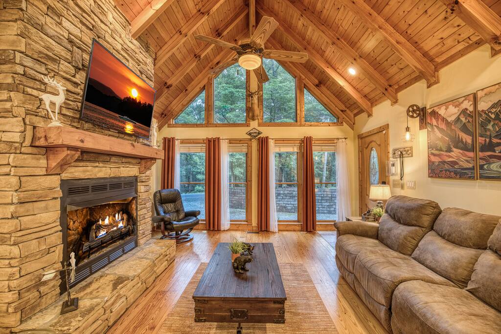 Cozy living room with a gas fireplace, large scenic window, and big-screen smart TV. Plush furnishings and earthy decor create a warm, inviting retreat. Cozy living room with a gas fireplace, large scenic window, and big-screen smart TV. Plush furnishings and earthy decor create a warm, inviting retreat. - Spacious Cabin, Hot Tub, Theater, Dog Run, FirePit - Ellijay