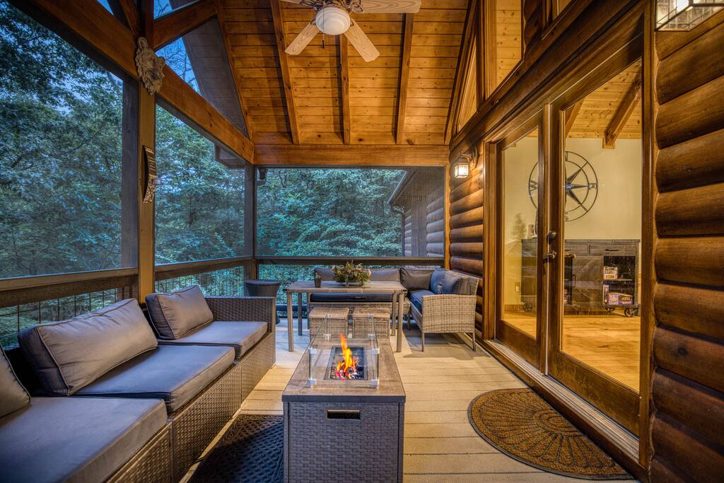Serene screened patio with a gas fire pit, featuring wicker seating and scenic woodland views. Wood beams and soft lighting add a rustic, cozy touch to the space. Serene screened patio with a gas fire pit, featuring wicker seating and scenic woodland views. Wood beams and soft lighting add a rustic, cozy touch to the space. - Spacious Cabin, Hot Tub, Theater, Dog Run, FirePit - Ellijay