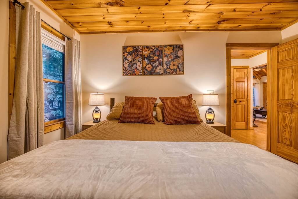 Cozy guest bedroom with a queen bed, plush bedding, and warm wood ceiling. Vibrant floral art, bedside lamps, and a scenic window with soft curtains create a relaxing vibe. Cozy guest bedroom with a queen bed, plush bedding, and warm wood ceiling. Vibrant floral art, bedside lamps, and a scenic window with soft curtains create a relaxing vibe. - Spacious Cabin, Hot Tub, Theater, Dog Run, FirePit - Ellijay