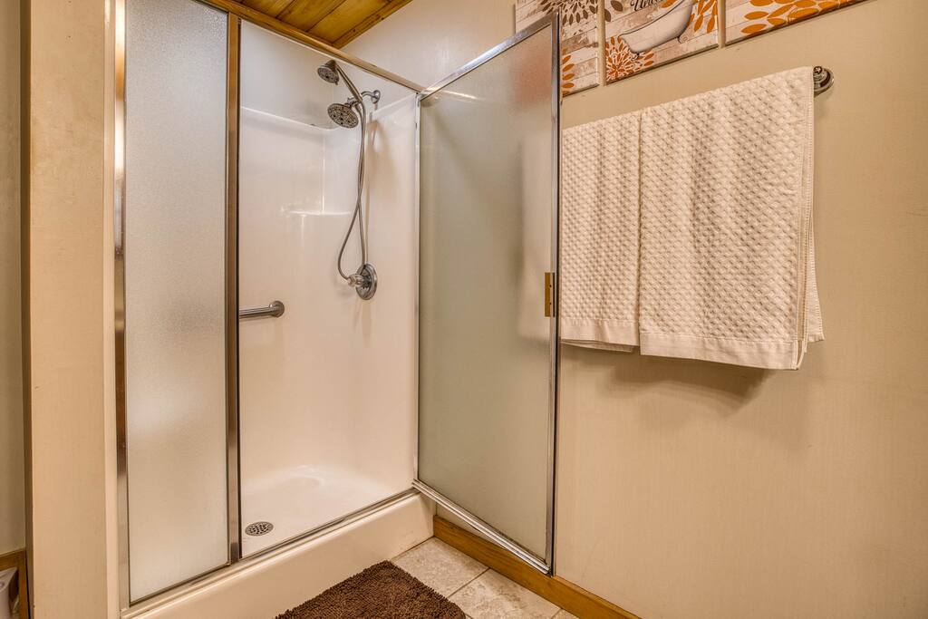 The master bathroom features a sleek glass-enclosed shower with dual showerheads, a modern design, and a cozy mat. Warm wood ceiling and decorative art add a rustic touch. The master bathroom features a sleek glass-enclosed shower with dual showerheads, a modern design, and a cozy mat. Warm wood ceiling and decorative art add a rustic touch. - Spacious Cabin, Hot Tub, Theater, Dog Run, FirePit - Ellijay