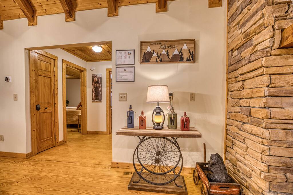 Warm hallway with wooden beams, stone wall, and hardwood floors. A rustic table with a lantern and "HOME" bottles sits under mountain art, complemented by a bear statue and cozy decor. Warm hallway with wooden beams, stone wall, and hardwood floors. A rustic table with a lantern and "HOME" bottles sits under mountain art, complemented by a bear statue and cozy decor. - Spacious Cabin, Hot Tub, Theater, Dog Run, FirePit - Ellijay