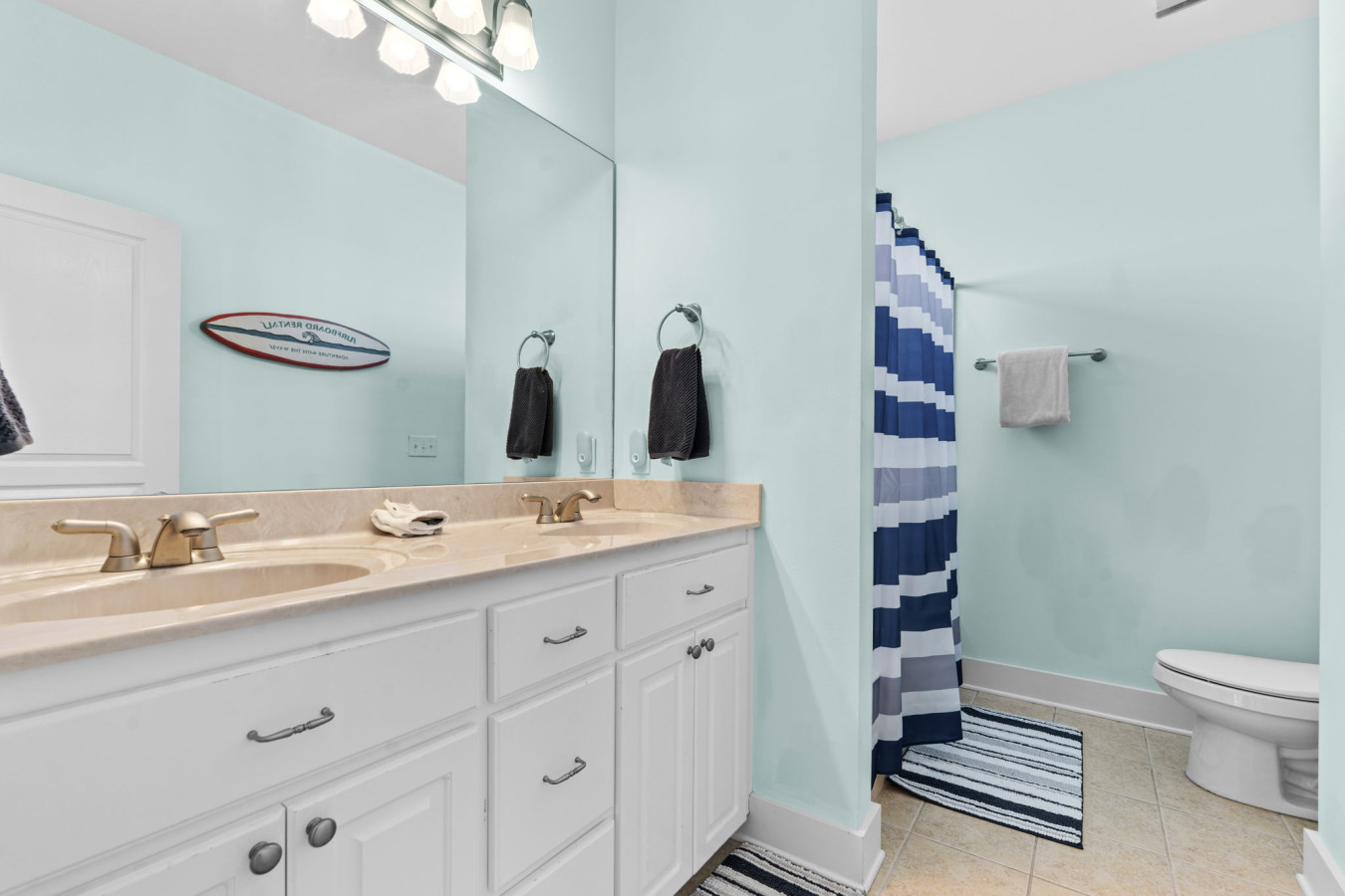 This full bathroom features double sinks, a walk-in shower with a bold blue striped curtain, and fun surf-themed decor.