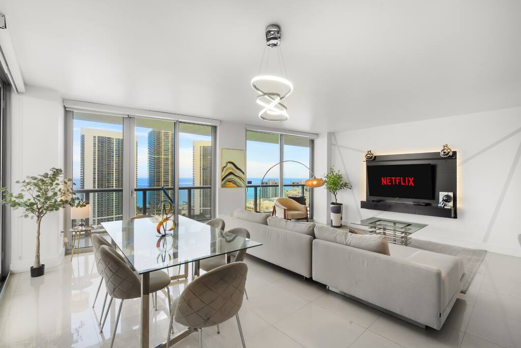 The dining area features a sleek glass table with seating for six, framed by floor-to-ceiling windows that bring in stunning ocean and city views.