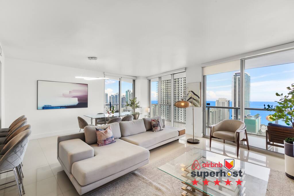 Surrounded by floor-to-ceiling windows, the living area offers designer décor, a comfortable couch, and direct views of the ocean. A modern dining table completes the space, ideal for gatherings day or night.