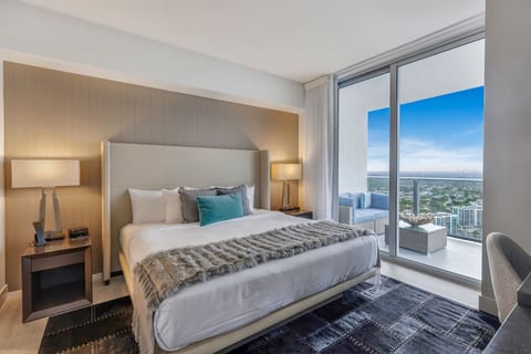 Step into the second bedroom, where a king-size bed, elegant lighting, and cozy design set the scene. An in-room bathroom and dedicated workspace add convenience, while sliding doors open to a balcony with sweeping city and ocean views.