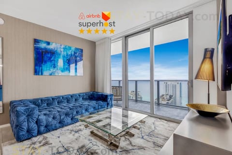 A view of the sparkling ocean sets the scene for this stylish living room featuring a deep blue velvet sofa and a sleek glass coffee table. The large sliding doors open to a balcony, inviting natural light and the refreshing sea breeze inside.