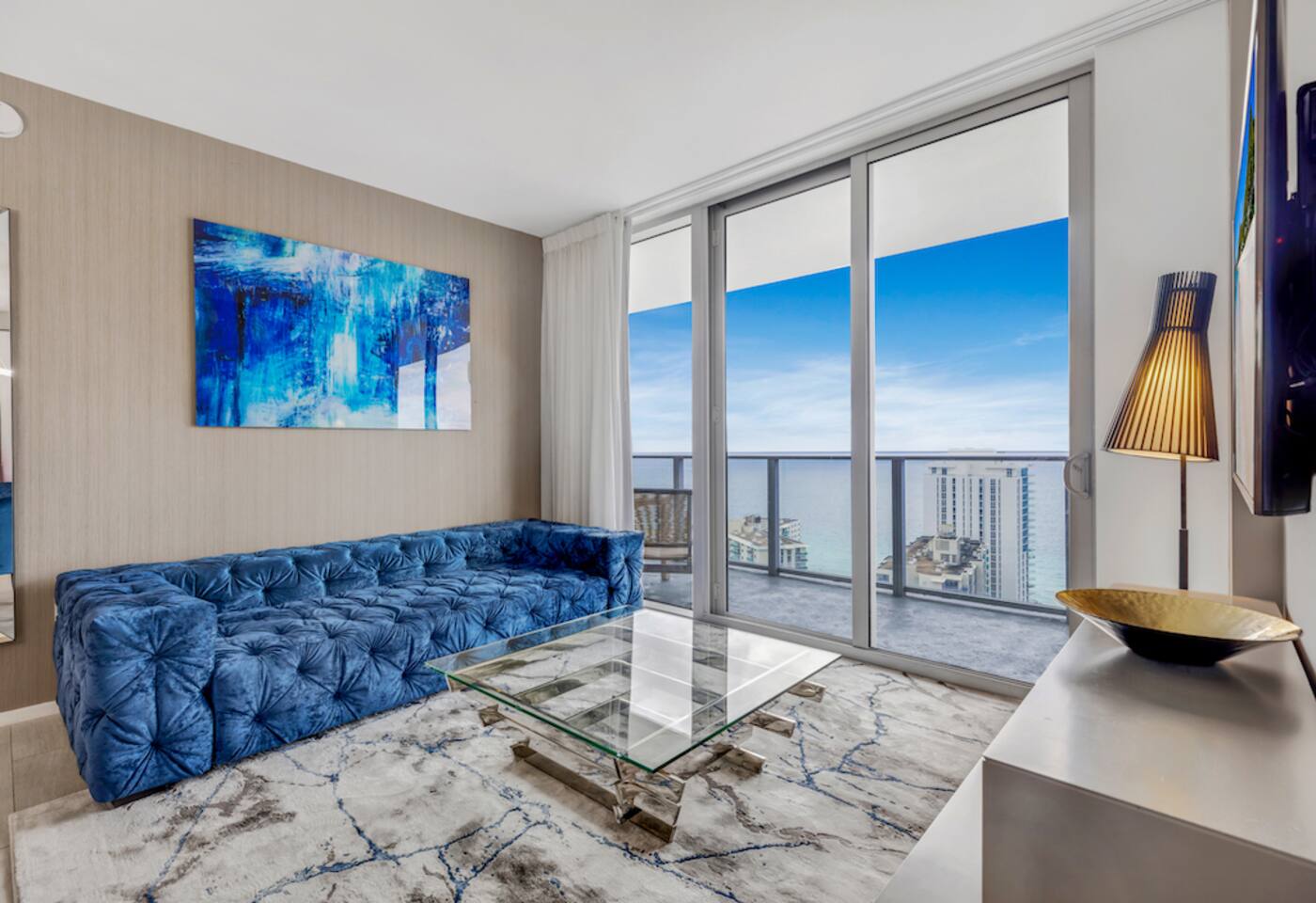 A view of the sparkling ocean sets the scene for this stylish living room featuring a deep blue velvet sofa and a sleek glass coffee table. The large sliding doors open to a balcony, inviting natural light and the refreshing sea breeze inside.