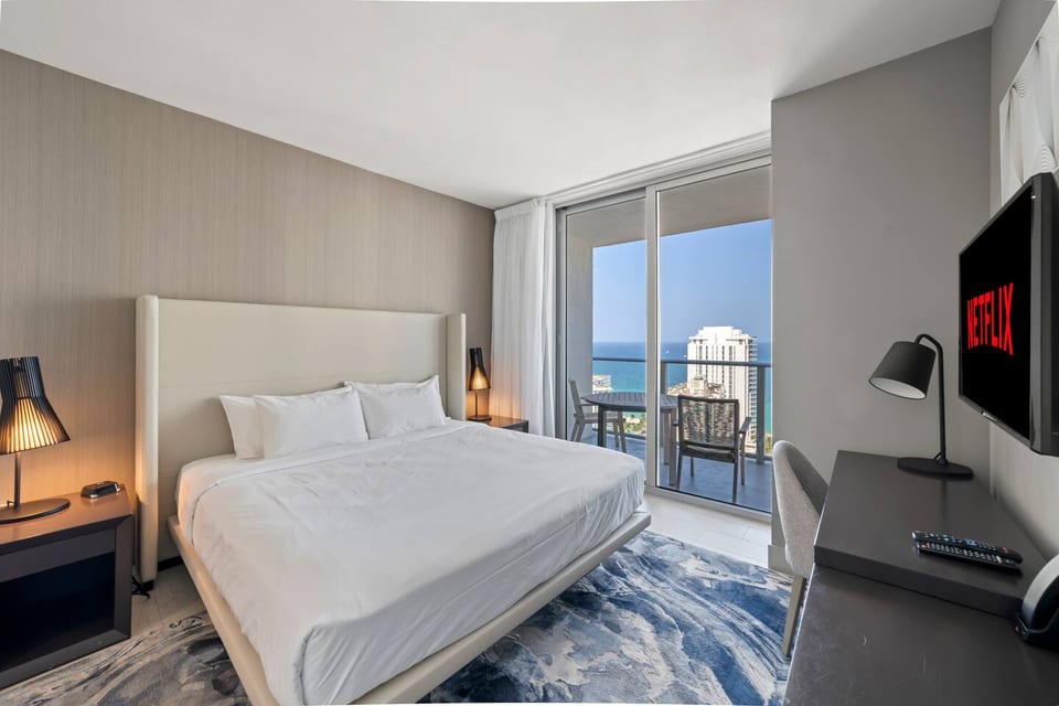 Step into a spacious master bedroom with a king-size bed, bedside tables, and a desk with a convenient lamp. The room opens onto a balcony with seating and ocean views, while the private en-suite bathroom adds comfort and convenience.